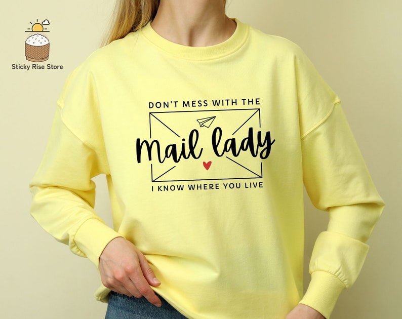 May include: Yellow long-sleeve sweatshirt with a black graphic print of an envelope with a paper airplane and the text "Don't mess with the mail lady I know where you live".