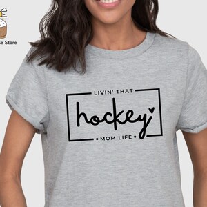 Może przedstawiać: Szara koszulka z czarnym nadrukiem "Livin' that hockey Mom Life".