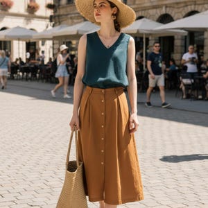 May include: A woman in a teal top and a brown button-down skirt, with a straw hat and a woven tote bag. The outfit is suitable for a sunny day. The background shows a European city with outdoor seating.