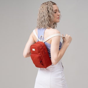 May include: A red canvas mini backpack with white straps. The backpack has a zipped pocket on the front and a small pocket on the side.