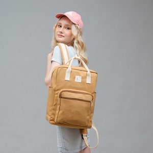 May include: A tan canvas backpack with a white zipper and a white strap. The backpack has a small pocket on the front. A person is wearing the backpack and a pink baseball cap.