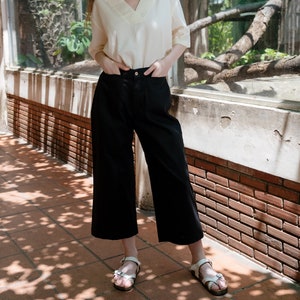 May include: A woman wearing a white v-neck top, black wide-leg trousers, and a straw hat. She is standing in front of a brick wall and a window with a green awning.
