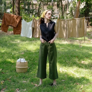 May include: A woman wearing a black sleeveless shirt, green trousers, and a yellow headscarf stands in a grassy area.  She is looking to the right of the frame.  There are several pieces of clothing hanging on a clothesline behind her.