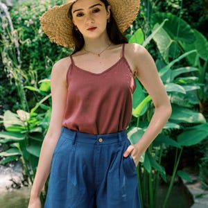 May include: A woman wearing a rust-colored tank top and blue denim shorts. She is wearing a wide-brimmed straw hat and has a necklace on.