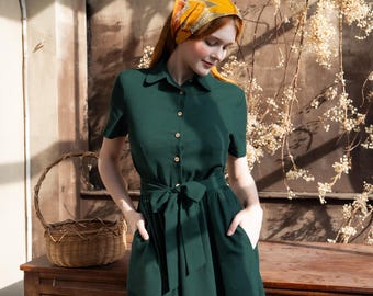 May include: A dark green dress with short sleeves, a collared neckline, and a tie waist. The dress has gold-colored buttons and pockets. A patterned yellow and orange headscarf is worn. A woven basket sits on a wooden table.