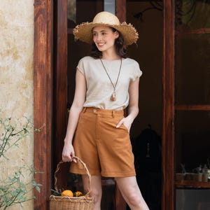May include: A woman in a straw hat, beige top, and rust-coloured shorts stands by a wooden door, holding a woven basket with oranges. She wears a long necklace with a pendant. The outfit suggests a casual, summery style, suitable for warm weather.