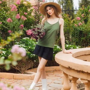 May include: A woman wearing a green tank top, black skirt, and white rain boots stands in a garden. She is holding a bouquet of purple flowers and wearing a straw hat.