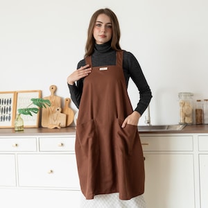 Peut inclure: Un tablier marron avec de grandes poches avant et une encolure carrée. Le tablier est porté sur un col roulé gris foncé. Il y a une petite étiquette blanche près de l'encolure. En arrière-plan, des planches à découper en bois et des œuvres encadrées.