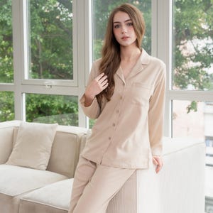 Op de afbeelding: Een beige pyjamaset. De set bestaat uit een overhemd met knopen en een borstzak en een bijpassende broek. De pyjama is gemaakt van een zachte, lichte stof. Het model draagt de pyjama in een goed verlichte kamer met een raam op de achtergrond.