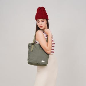 May include: A woman wearing a red knit beanie and a striped shirt is holding a green canvas tote bag with a strap.