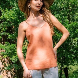 May include: A woman wearing a straw hat, a peach-colored tank top, and white shorts. She is holding a woven bag and standing in front of green foliage.