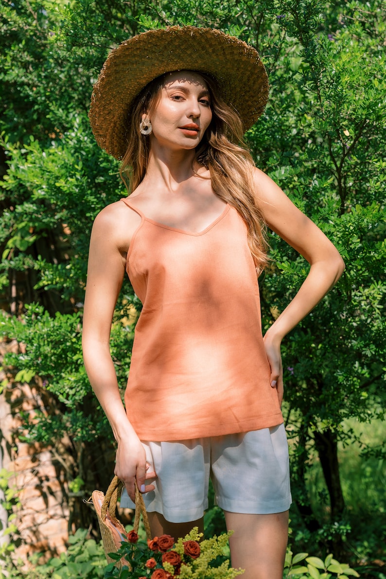 May include: A woman wearing a straw hat, a peach-colored tank top, and white shorts. She is holding a woven bag and standing in front of green foliage.