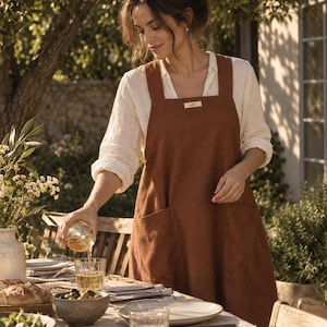 May include: A rust-coloured linen apron featuring wide shoulder straps, a square neckline, and two large front pockets. A small rectangular label with text is stitched onto the centre of the chest area. The apron is worn over a white long-sleeved shirt.