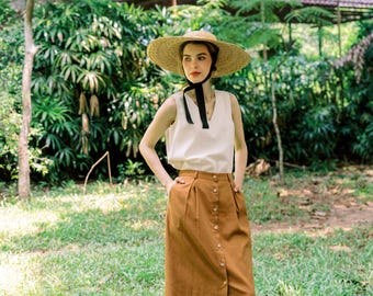 Peut inclure: Une femme portant un haut blanc sans manches et une jupe marron à boutons. Elle porte un chapeau de paille à larges bords avec un ruban noir. Elle se tient dans une zone herbeuse.