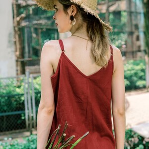 May include: A woman wearing a red tank top with a wide brimmed straw hat. She is holding a bundle of green branches in her right hand.