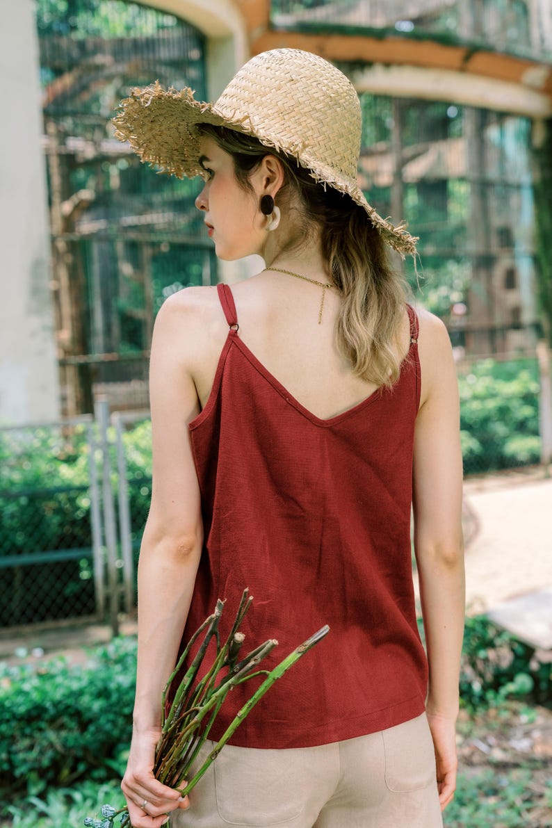 May include: A woman wearing a red tank top with a wide brimmed straw hat. She is holding a bundle of green branches in her right hand.