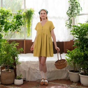 May include: A mustard yellow, short-sleeved dress with a V-neck and button details. The dress has a gathered waist and falls to mid-thigh. The person is wearing white socks and brown sandals, holding a small woven basket. The background features lush greenery.
