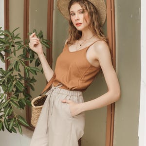 May include: A woman wearing a brown tank top, beige pants, and a wide-brimmed straw hat. She is standing next to a window and holding a wicker basket.