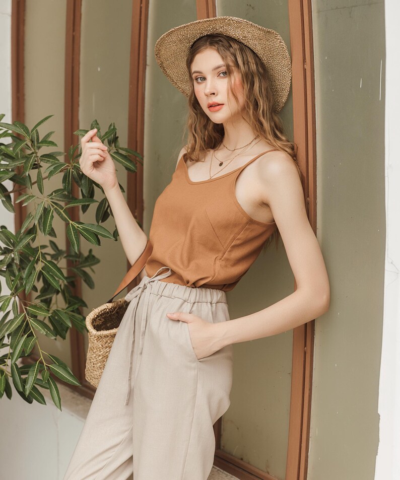 May include: A woman wearing a brown tank top, beige pants, and a wide-brimmed straw hat. She is standing next to a window and holding a wicker basket.