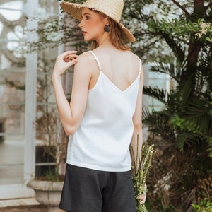 May include: A woman wearing a white tank top and black shorts, holding a bouquet of green stems. She is wearing a straw hat and has her hair pulled back.