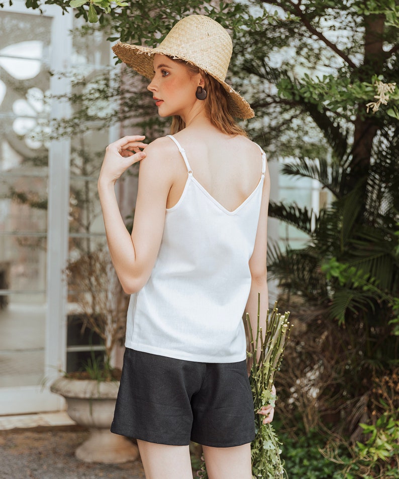 May include: A woman wearing a white tank top and black shorts, holding a bouquet of green stems. She is wearing a straw hat and has her hair pulled back.