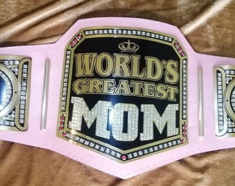 Könnte beinhalten: Ein rosa Meisterschaftsgürtel mit goldenen Akzenten und Strasssteinen. Die mittlere Platte hat einen schwarzen Hintergrund mit dem Text "WORLD'S GREATEST MOM" in goldenen Buchstaben. Die seitlichen Platten haben einen goldenen Hintergrund mit dem Text "BEST MOM EVER" in schwarzen Buchstaben.