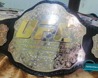 Könnte beinhalten: Ein gold- und silberner UFC-Championschaftsgürtel mit schwarzen Lederriemen. Der Gürtel trägt das UFC-Logo und die Worte "Ultimate Fighting Championship".