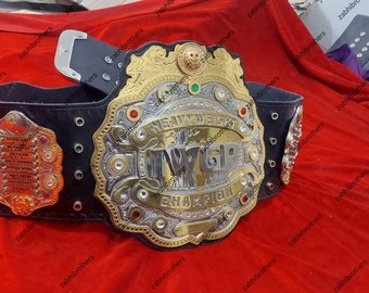 May include: A gold and silver championship belt with a black leather strap. The belt features a large, ornate center plate with the words "Heavyweight Champion" and "IWGP" in silver lettering. The belt is decorated with red and green jewels.