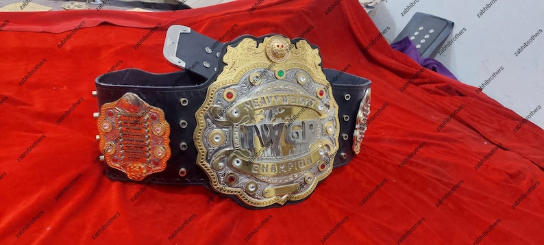K&ouml;nnte beinhalten: Ein gold- und silberner Meisterschaftsg&uuml;rtel mit einem schwarzen Lederriemen. Der G&uuml;rtel verf&uuml;gt &uuml;ber eine gro&szlig;e, verzierte Mittelplatte mit den Worten "Heavyweight Champion" und "IWGP" in silbernen Lettern. Der G&uuml;rtel ist mit roten und gr&uuml;nen Juwelen verziert.