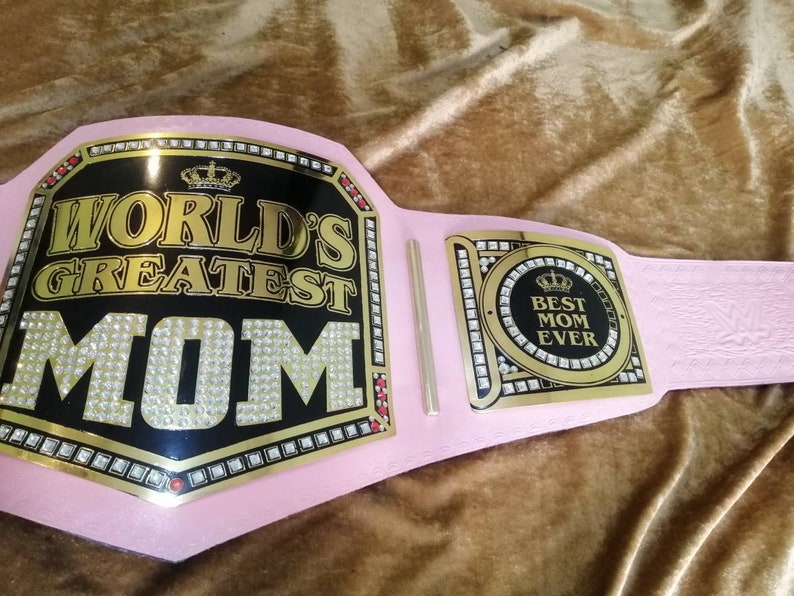May include: A pink championship belt with gold accents and rhinestones. The center plate reads "World's Greatest Mom" and the side plate reads "Best Mom Ever".