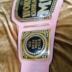 May include: Pink and gold championship belt with rhinestones. The belt has two plates. The top plate says "World's Greatest Mom" and the bottom plate says "Best Mom Ever".