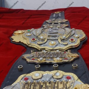 K&ouml;nnte beinhalten: Ein schwarzer Leder-Champion-G&uuml;rtel mit goldenen und silbernen Akzenten. Der G&uuml;rtel verf&uuml;gt &uuml;ber eine gro&szlig;e, verzierte Mittelplatte mit dem Wort "CHAMPION" in silbernen Buchstaben. Die Platte ist von kleineren Platten mit den Namen der ehemaligen Champions umgeben.