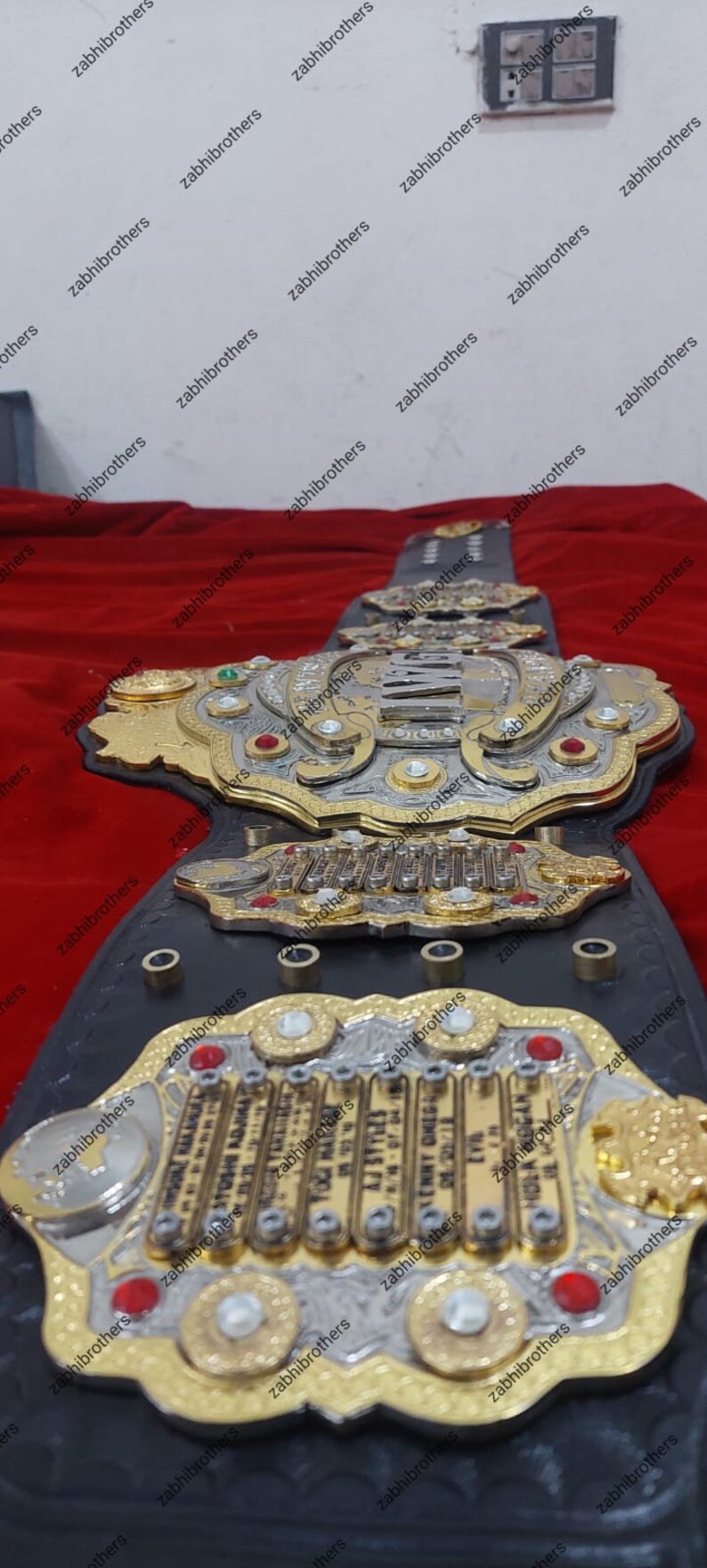 K&ouml;nnte beinhalten: Ein schwarzer Leder-Champion-G&uuml;rtel mit goldenen und silbernen Akzenten. Der G&uuml;rtel verf&uuml;gt &uuml;ber eine gro&szlig;e, verzierte Mittelplatte mit dem Wort "CHAMPION" in silbernen Buchstaben. Die Platte ist von kleineren Platten mit den Namen der ehemaligen Champions umgeben.
