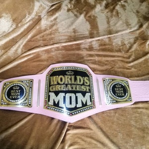 May include: Pink championship belt with gold accents and rhinestones. The center plate reads "World's Greatest Mom" with a crown above the text. The side plates read "Best Mom Ever".