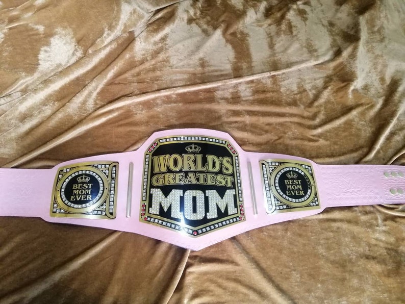 May include: Pink championship belt with gold accents and rhinestones. The center plate reads "World's Greatest Mom" with a crown above the text. The side plates read "Best Mom Ever".