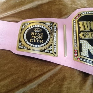 May include: Pink and gold championship belt with rhinestones and the text "World's Greatest Mom" and a smaller plaque with the text "Best Mom Ever" and a crown.