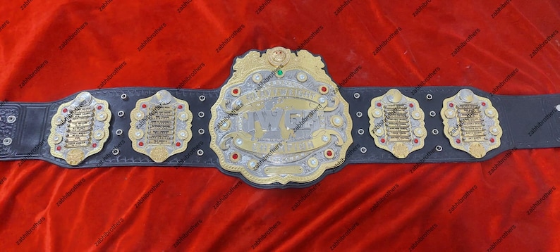 K&ouml;nnte beinhalten: Ein schwarzer Leder-Wrestling-Champion-G&uuml;rtel mit goldenen und silbernen Akzenten. Die Mittelplatte zeigt den Text "International Wrestling Federation" und "IWF" in einer stilisierten Schriftart. Der G&uuml;rtel hat vier Seitenplatten mit goldenen und silbernen Akzenten.