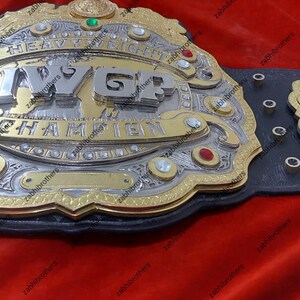 K&ouml;nnte beinhalten: Ein gold- und silberner Meisterschaftsg&uuml;rtel mit der Aufschrift "IWGP Heavyweight Champion" auf der Vorderseite. Der G&uuml;rtel hat ein schwarzes Lederband mit silbernen Nieten und eine gro&szlig;e, verzierte Mittelplatte.