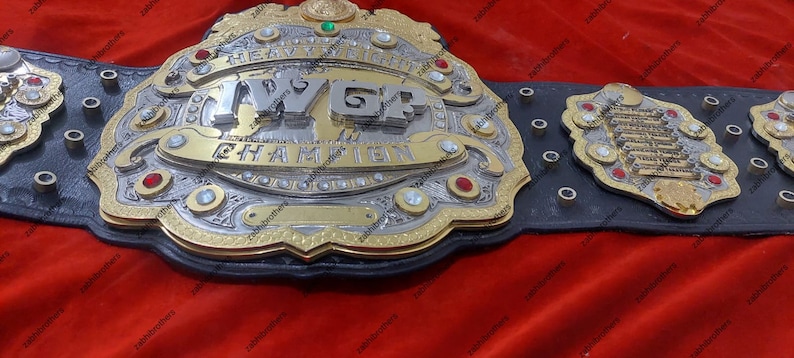 K&ouml;nnte beinhalten: Ein gold- und silberner Meisterschaftsg&uuml;rtel mit der Aufschrift "IWGP Heavyweight Champion" auf der Vorderseite. Der G&uuml;rtel hat ein schwarzes Lederband mit silbernen Nieten und eine gro&szlig;e, verzierte Mittelplatte.