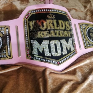 May include: A pink championship belt with gold accents and rhinestones. The center plate features a black background with gold lettering that reads "World's Greatest Mom". The side plates feature a crown and the words "Best Mom Ever".