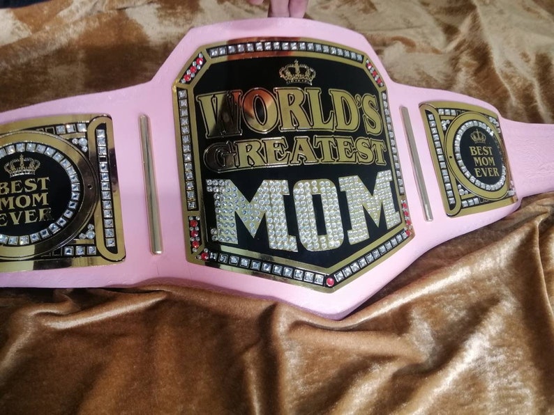 May include: A pink championship belt with gold accents and rhinestones. The center plate features a black background with gold lettering that reads "World's Greatest Mom". The side plates feature a gold crown and the text "Best Mom Ever".