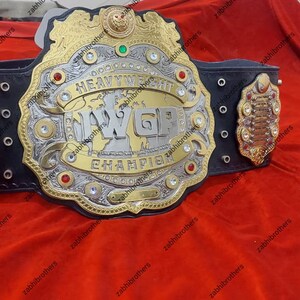 K&ouml;nnte beinhalten: Ein gold- und silberner Wrestling-Championschaftsg&uuml;rtel mit einem schwarzen Lederriemen. Der G&uuml;rtel verf&uuml;gt &uuml;ber eine gro&szlig;e Mittelplatte mit den Worten "Weltmeister im Schwergewicht" und den Buchstaben "WGP" in einer stilisierten Schrift. Der G&uuml;rtel ist mit Strasssteinen und anderen Verzierungen verziert.