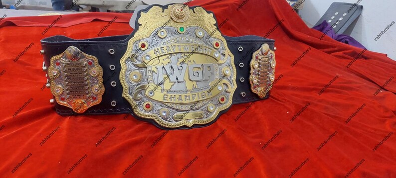 K&ouml;nnte beinhalten: Ein gold- und silberner Wrestling-Championschaftsg&uuml;rtel mit einem schwarzen Lederriemen. Der G&uuml;rtel verf&uuml;gt &uuml;ber eine gro&szlig;e Mittelplatte mit den Worten "Weltmeister im Schwergewicht" und den Buchstaben "WGP" in einer stilisierten Schrift. Der G&uuml;rtel ist mit Strasssteinen und anderen Verzierungen verziert.