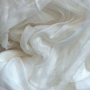 Pode incluir: Tecido branco transparente com uma trama texturizada, drapeado e dobrado para criar um visual suave e fluido.