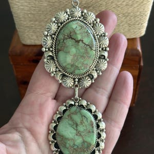 May include: A silver pendant necklace featuring two large, oval-shaped turquoise stones. Each stone is set in a decorative silver frame with a rope-like border and a floral design. The necklace is displayed against a neutral background.