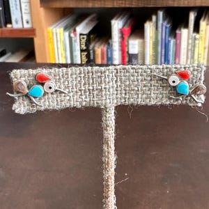 Könnte beinhalten: Ein Paar silberfarbene Ohrstecker in Vogelform. Jeder Ohrring hat einen roten, weißen und türkisfarbenen Stein mit Drahtdetails für Flügel und Schwanz. Die Ohrringe werden auf einem strukturierten, beigefarbenen Stoff präsentiert.