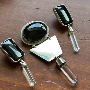 Könnte beinhalten: Eine Sammlung von silberfarbenen Ohrringen und einem Anhänger. Die Ohrringe haben schwarze, rechteckige Steine und Anhänger aus klarem Kristall. Der Anhänger hat einen großen ovalen schwarzen Stein, eine silberne geometrische Form und einen Anhänger aus klarem Kristall.