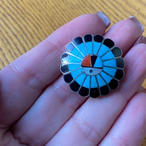 May include: A round, silver-toned pendant with a turquoise, black, white, and orange sunburst design. The pendant is held in a hand, showcasing the intricate details of the inlaid stones. The design is reminiscent of Native American jewelry.