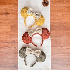 May include: Three handmade crochet headbands with mouse ears and a bow. The headbands are in mustard yellow, rust, and olive green. The bows are a neutral beige color. The headbands are displayed on a white runner with pinecones and a decorative pumpkin.