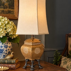 May include: A table lamp featuring a rectangular off-white fabric shade atop a metallic gold-toned, four-sided base with a textured finish and white dotted patterns. The base is supported by four dark, ornate, claw-style metal feet.